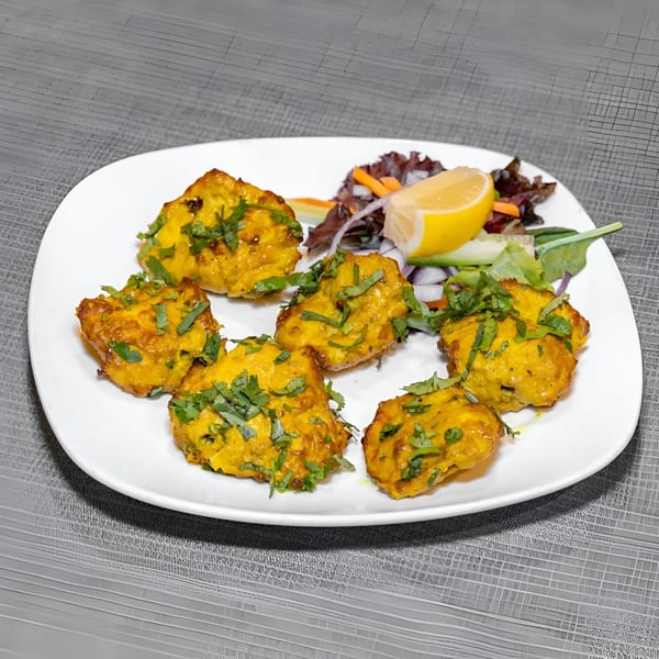 Six pieces of golden Indian chicken tikka with cilantro, a lemon wedge, and salad on a white plate.