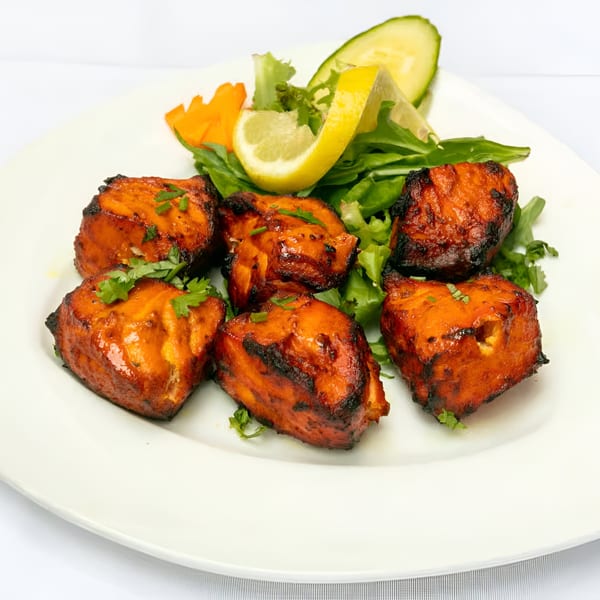 Six pieces of Indian chicken tikka, garnished with cilantro, lemon, and salad on a white plate.